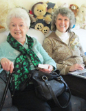 Two smiling women sitting on a couch together with several teddy bears visible in the background.