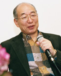 Hiroshi Doi speaking into a microphone, wearing glasses and a patterned shirt under a dark jacket.