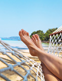 A first-person view of a person’s feet resting in a white rope hammock on a sunny tropical beach.