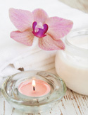 A calming spa arrangement with a lit pink candle, a pink orchid, and a jar of cream on a white towel.