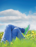 Low-angle view of a person wearing blue jeans and green sneakers relaxing in a field of yellow wildflowers.