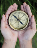 Close-up of two hands cupping a vintage brass compass against a blurred green grass background.