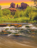 A scenic landscape featuring a flowing river in the foreground and red rock buttes under an orange sunset sky.