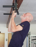 A senior man with a white beard performing a pull-up on a black multi-grip bar indoors.