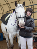 A smiling woman in a dark puffer jacket stands next to a large white horse in an indoor arena.