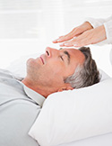 A man lies with eyes closed while a practitioner holds their hands just above his forehead for a Reiki session.