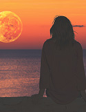 Silhouette of a woman sitting by the ocean, watching a massive, glowing orange moon rise over the horizon.