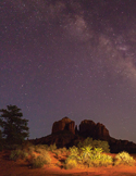 A night photo of the Milky Way galaxy over the silhouetted red rock formations of Sedona, Arizona.