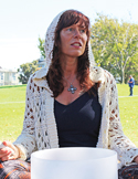 A woman in a hooded crochet cardigan sits outdoors behind a large white crystal singing bowl.