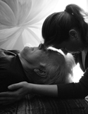 Black and white photo of a woman leaning down to touch foreheads with an elderly person lying in bed.