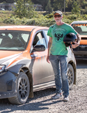 A man in a green shirt and sunglasses holding a helmet while standing next to an orange rally car.