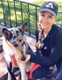 A smiling woman in a black baseball cap kneeling next to a speckled blue merle collie dog outdoors.
