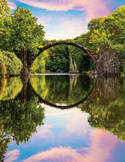 A stone arch bridge reflecting in a still river, forming a perfect circle surrounded by lush green trees.