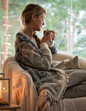 A woman wrapped in a cozy blanket sits in a chair, holding a warm mug in a dimly lit, peaceful room.