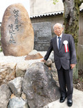 An elderly man in a suit stands next to a large stone memorial with Japanese calligraphy.