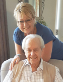 A smiling woman leaning over the shoulder of an elderly man, both looking at the camera indoors.