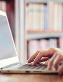 Close-up of hands typing on a laptop keyboard with a blurred bookshelf in the background.