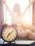 An alarm clock in the foreground with a blurred person stretching in front of a sunny window.