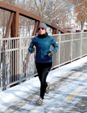 A person dressed in winter athletic gear runs across a snowy bridge on a bright day.