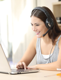 A smiling woman wearing a headset while working on a laptop in a bright room.
