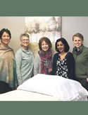 Five women smiling together in a clinical setting behind a white treatment table.