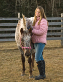 A young woman in a pink puffer jacket gently petting a speckled horse in a fenced paddock.