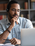 A man wearing a headset and glasses looks thoughtfully at his laptop screen while holding a pen.