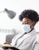 A woman wearing a medical face mask sits indoors while reading a book.