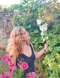A woman with curly hair stands in a lush garden admiring tall white and pink flowers.