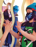 A child in a face mask and beanie holds their hands up toward a mirror reflection.