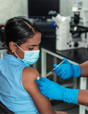 A healthcare worker in blue gloves administering a vaccine to a woman's arm in a medical setting.
