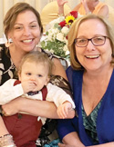 Two smiling women pose closely together, with one holding a young toddler in a vest and bowtie.