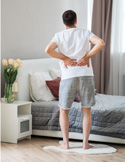 A man standing in a bedroom holding his lower back in pain, suggesting a back injury.