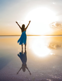 Woman in a blue dress with arms raised standing on a reflective salt flat at sunset.