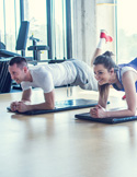 A man and woman smiling while holding a plank position on mats in a bright gym.
