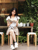 A young woman sitting outside on a stool, focused on reading a book amidst lush green plants.