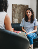 A woman in a blue shirt sits on a sofa, listening intently during a therapy or coaching session.