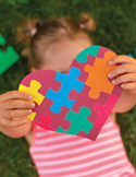 A young girl on green grass holds a heart-shaped puzzle made of colorful interlocking pieces over her face.