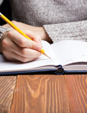 A close-up view of a hand writing in a white notebook with a yellow pencil on a wooden desk.