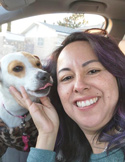 A smiling woman holds a small dog's chin as it licks her face while sitting in a car.