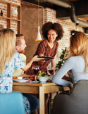 A woman stands and smiles while talking to a group of people seated at a cafe table.