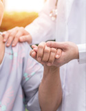 A healthcare worker in a white coat comfortingly holds the hand and shoulder of an elderly patient.