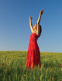 A woman in a red dress stands in a green field with arms raised, celebrating freedom beneath a clear blue sky.