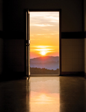 An open door reveals a glowing sunrise over distant hills, reflected on a polished floor in a dark interior.