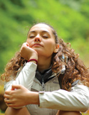 A woman with curly hair sits peacefully in nature with her eyes closed, appearing calm and meditative.