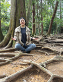 A woman meditates in a cross-legged pose amidst the large, sprawling surface roots of trees in a forest.