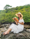 A woman in a white dress and sun hat sits on coastal rocks reading a book in front of lush green trees.