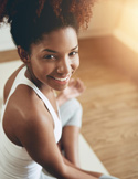 A smiling woman with dark curly hair sits on a yoga mat in a sunlit room, looking up at the camera.