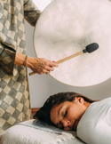 Person resting on a massage table while someone holds a large drum and mallet above them during a calming healing session.