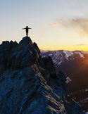 Person standing on a rocky mountain peak at sunset with arms outstretched, overlooking snow-covered ridges and glowing sky.